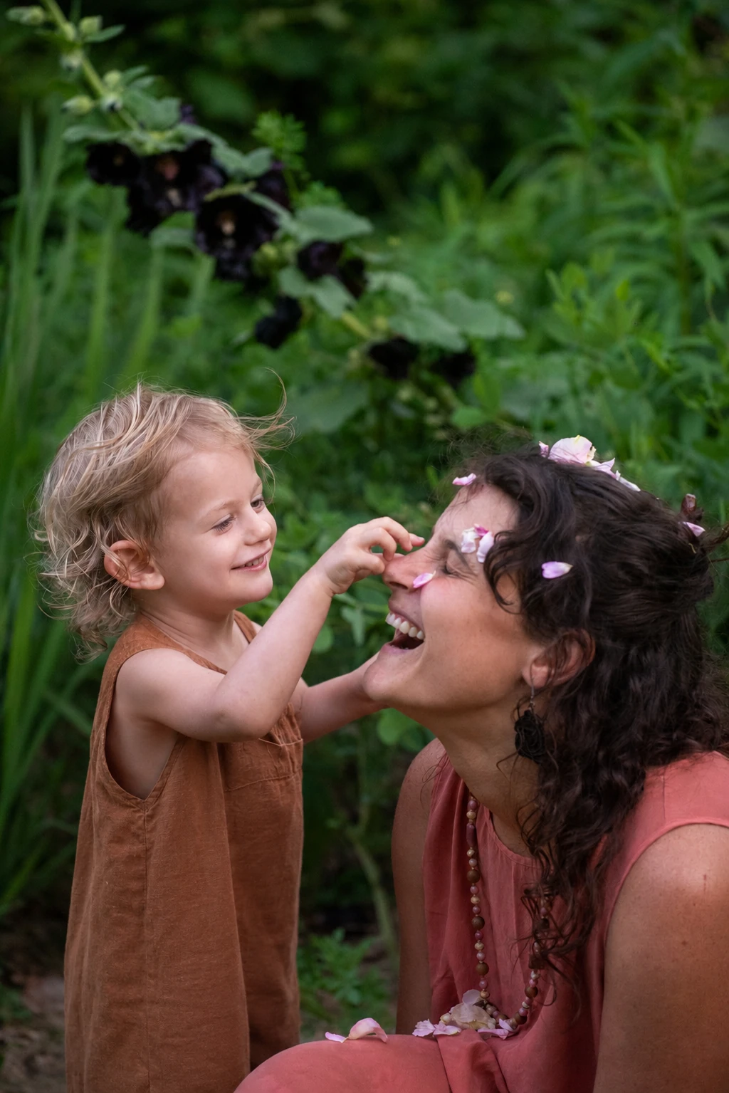 Laughing mother with her eyes closed as her toddler gently touches a flower petal to her nose in a lush Asheville garden